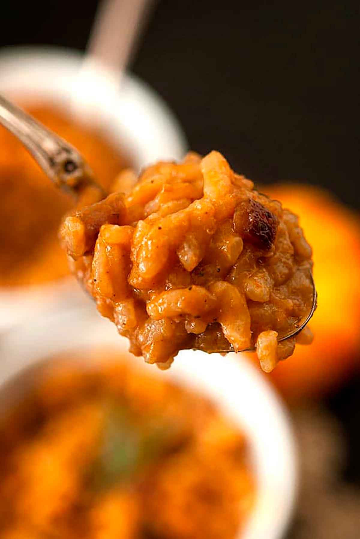 A spoon of pumpkin risotto held above bowls of risotto and a small pumpkin.