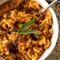 A bowl of pumpkin risotto, garnished with 2 sage leaves, on a lace table cloth beside another bowl and small pumpkin.