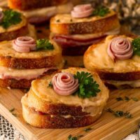 6 mini croque monsieur with a small ham rose garnishes and parsley on a wooden cutting board, surrounded by flowers.