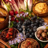 A crudités platter with vegetables, dips, olives, grapes and a bowl of bread on a tray.