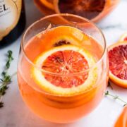 A blood orange champagne cocktail, garnished with an orange slice, beside a few stray orange slices on a table.