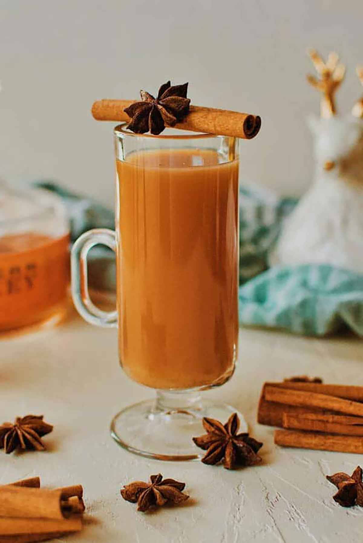 A cocktail in a mug, garnished with cinnamon, anise, and surrounded by cinnamon sticks and anise on a table.