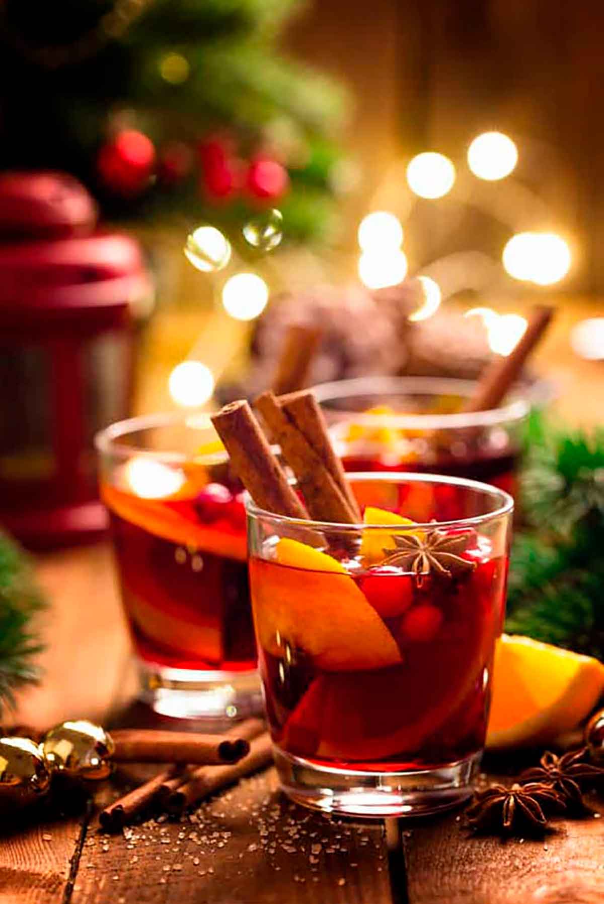 3 Gluhwein cocktails, garnished with cinnamon, orange and cranberries in a holiday setting.