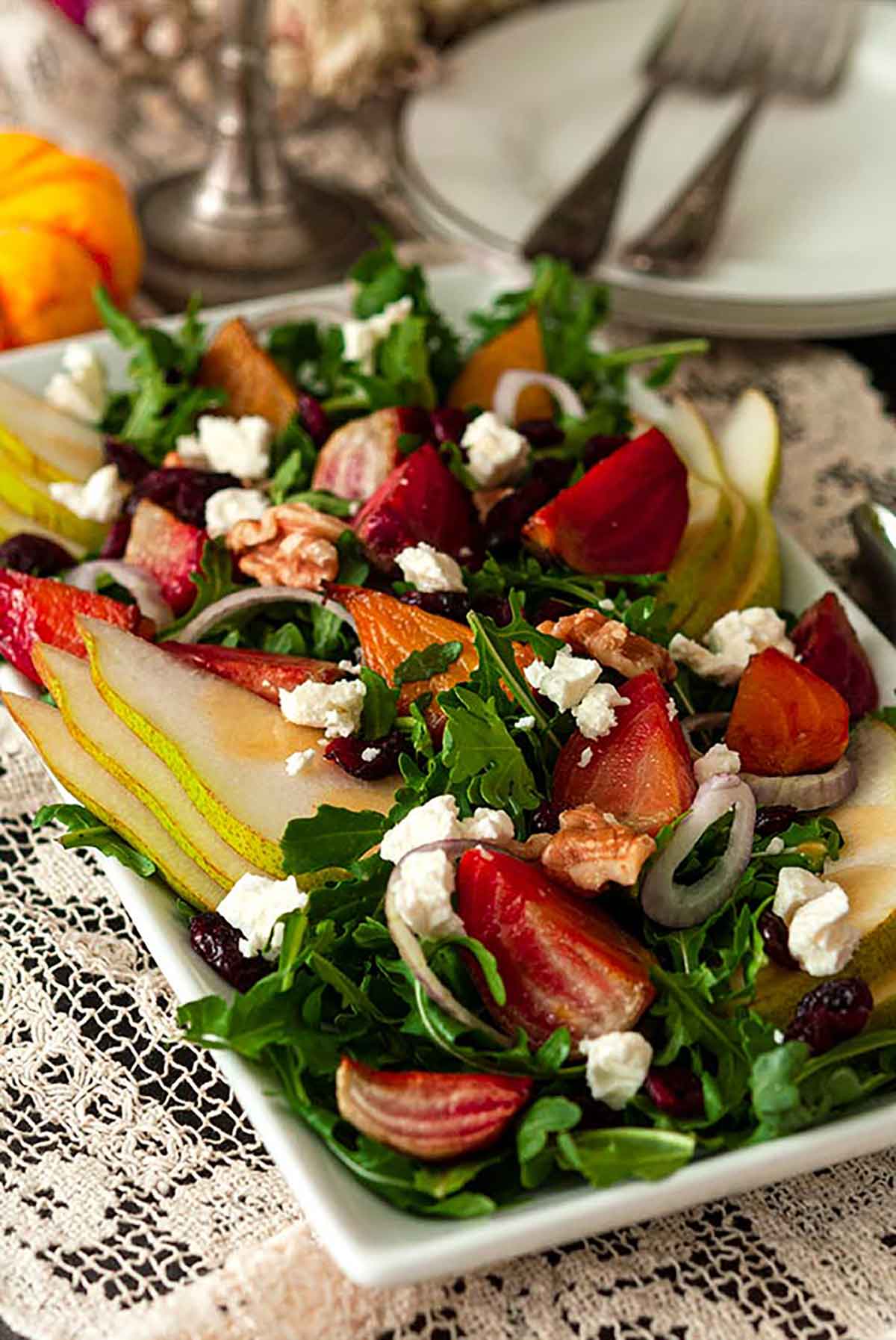 A salad with colorful vegetables, pears, berries and nuts on a lace table cloth with plates and forks in the background.