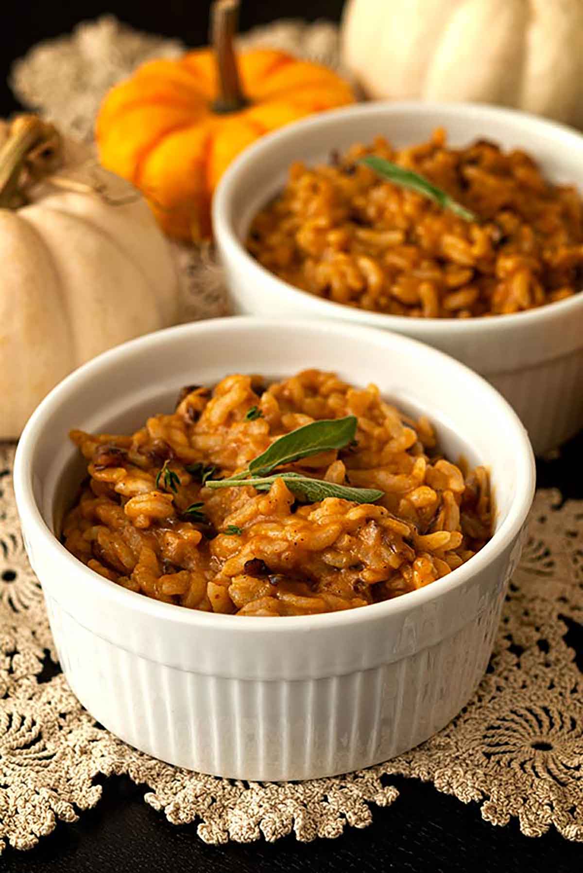 2 bowls of pumpkin risotto, garnished with sage leaves, on a lace table cloth with 3 small pumpkins in the background.