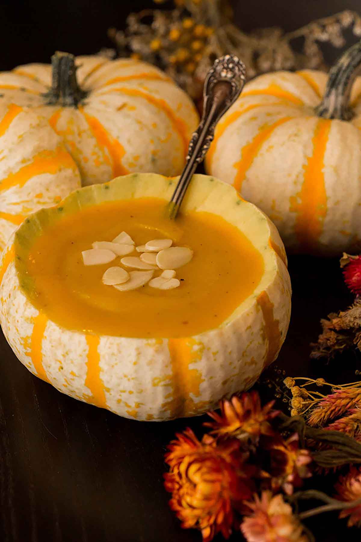 A pumpkin gourd full of soup, garnished with almonds, surrounded by dry flowers and more pumpkin gourds on a black table.