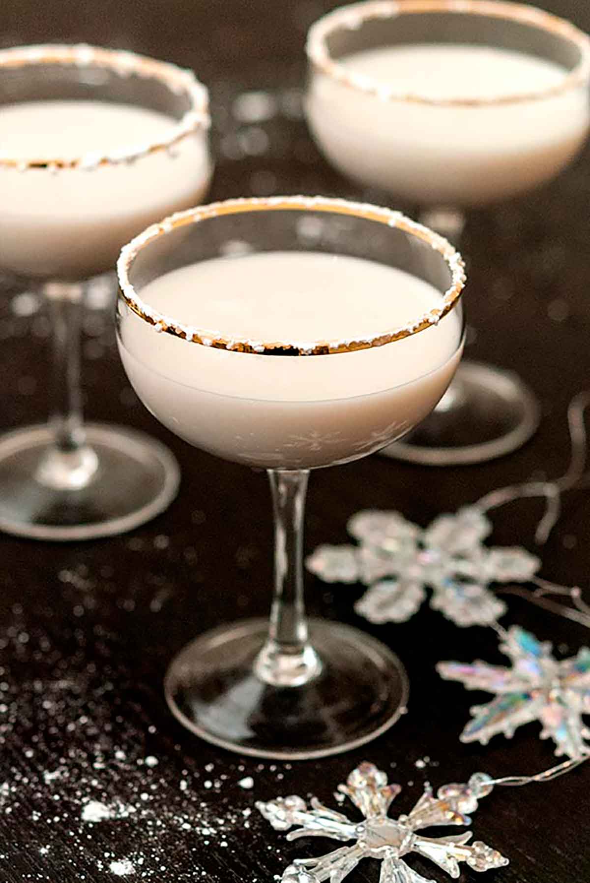 3 white cocktails, rimmed with powdered sugar, on a black table, sprinkled with sugar and with a few snowflake ornaments.