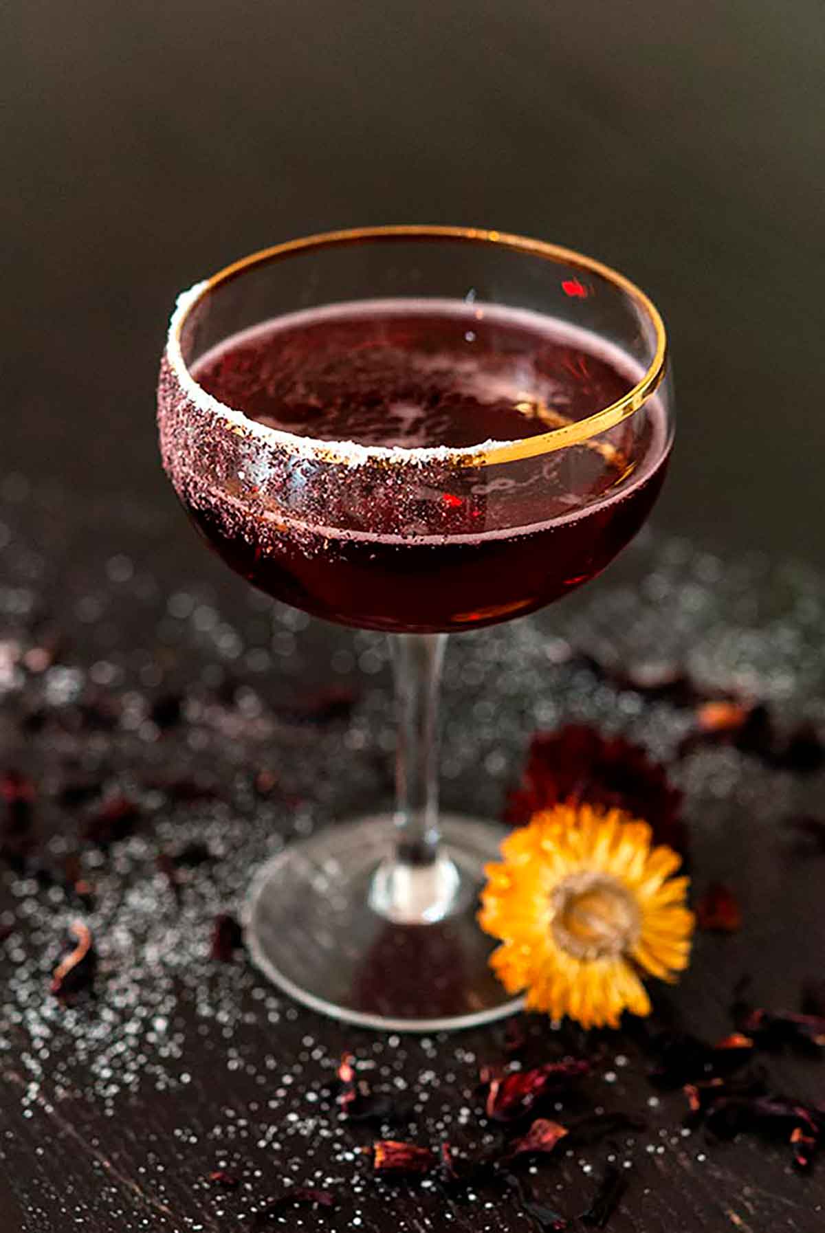 A sparkling spiced hibiscus cocktail on a dark table, sprinkled with sugar and cloves, with a tiny flower placed beside it.