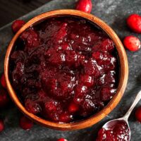 A bowl of cranberry sauce on a stone slate, surrounded by a few fresh cranberries, and a spoon of sauce.