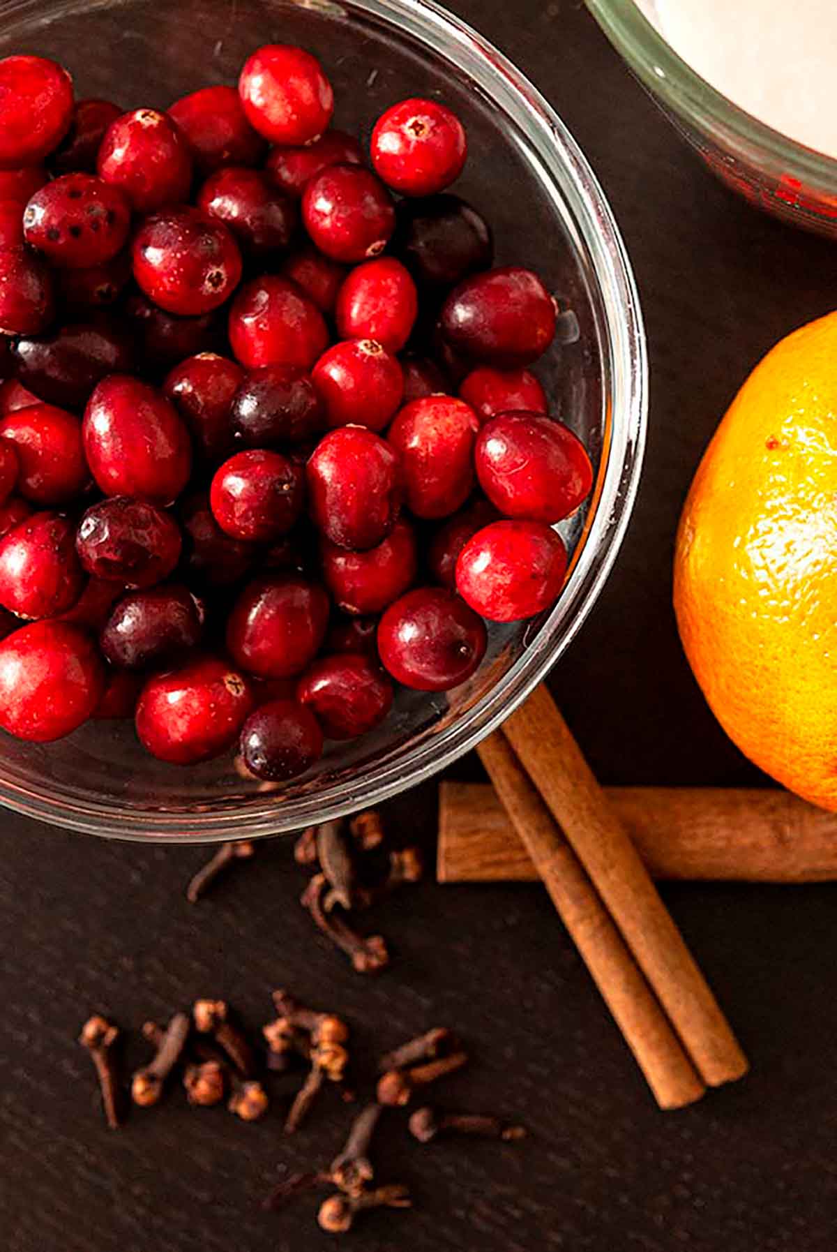 A bowl of cranberries beside 2 cinnamon sicks, a sprinkling of cloves and an orange on a table.