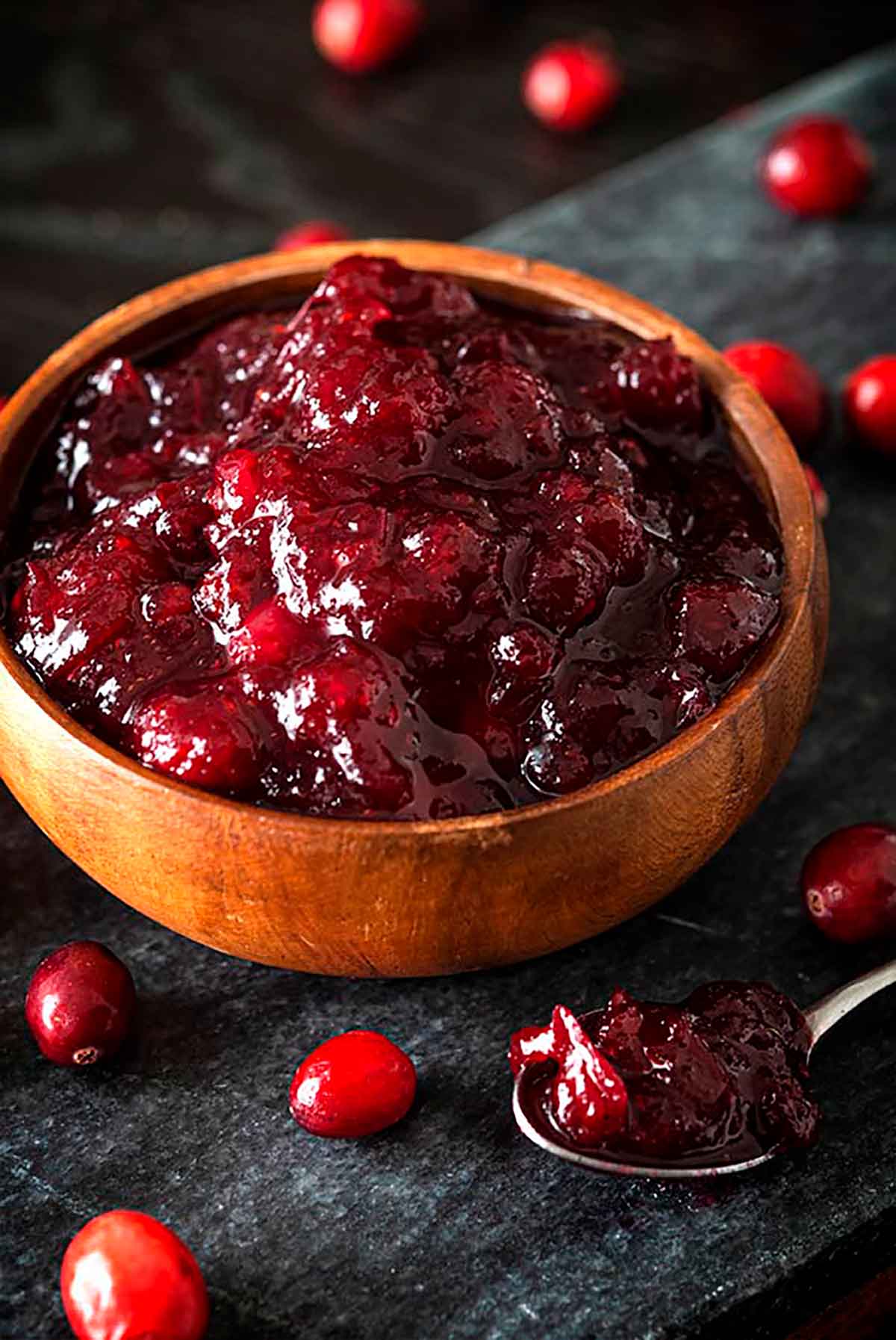 A bowl of cranberry sauce on a stone slate, surrounded by a few fresh cranberries, and a spoon of sauce.