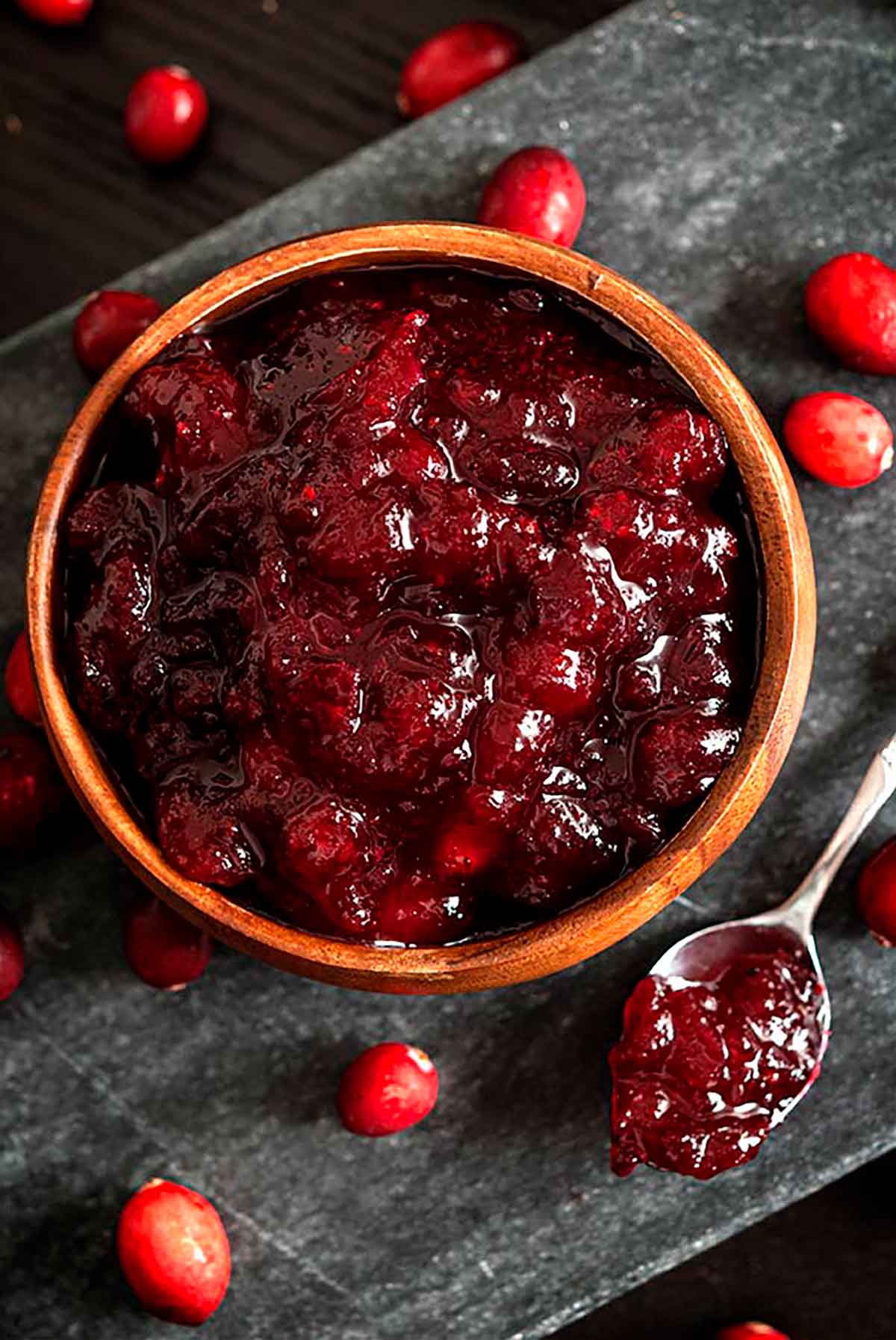 A bowl of cranberry sauce on a stone slate, surrounded by a few fresh cranberries, and a spoon of sauce.
