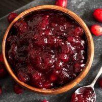 A bowl of cranberry sauce on a stone slate, surrounded by a few fresh cranberries, and a spoon of sauce.
