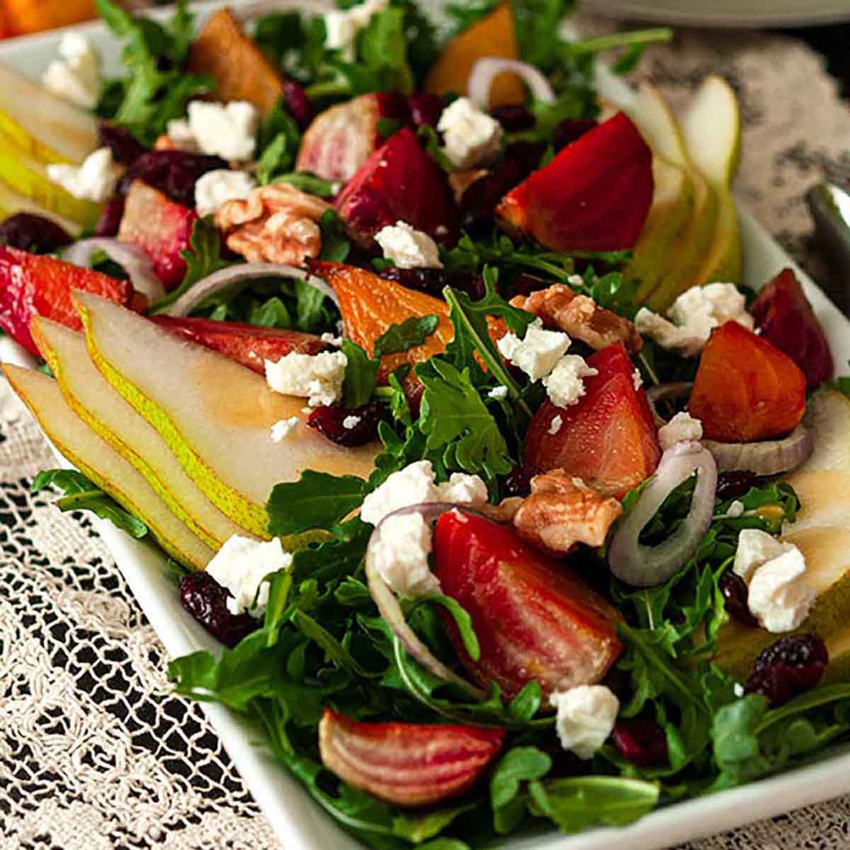 A salad with colorful vegetables, pears, berries and nuts on a lace table cloth with plates and forks in the background.