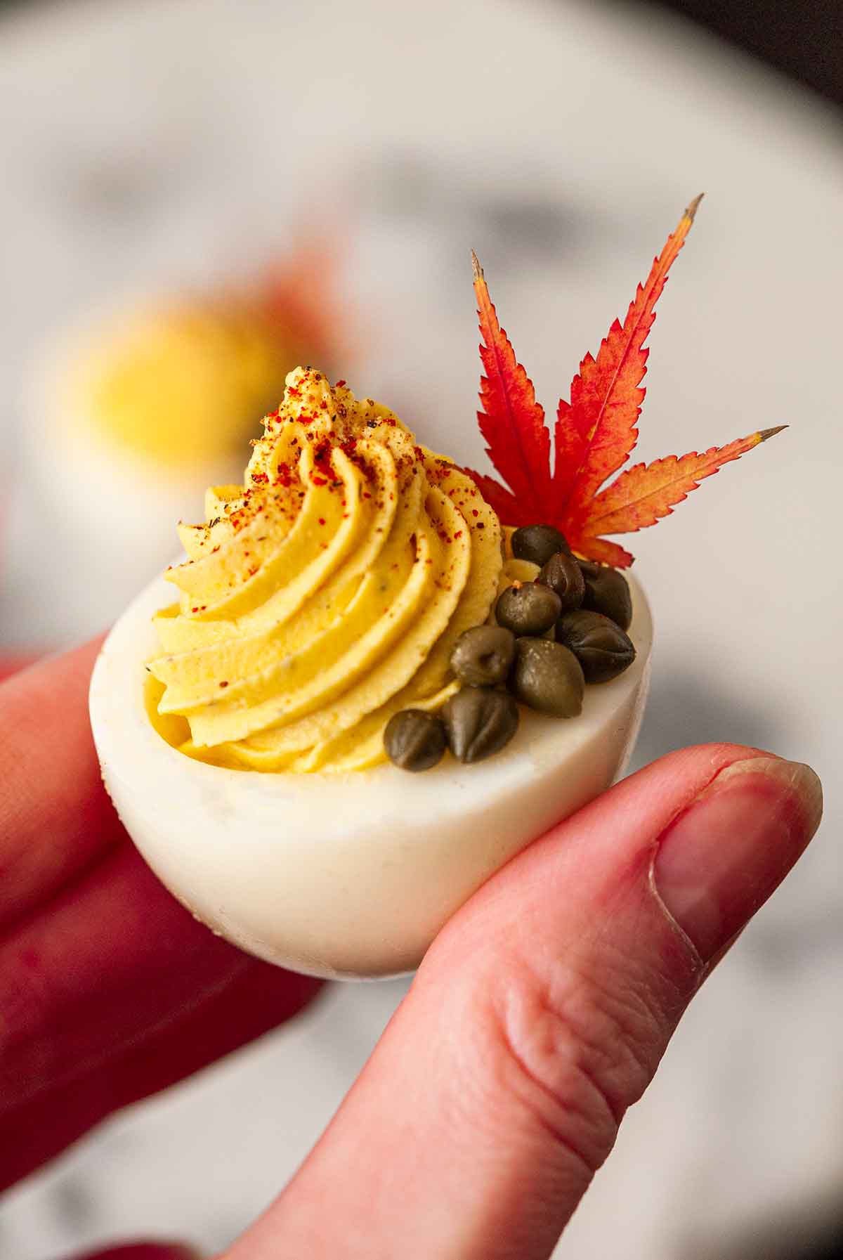 Fingers holding a deviled egg garnished with capers and a leaf.