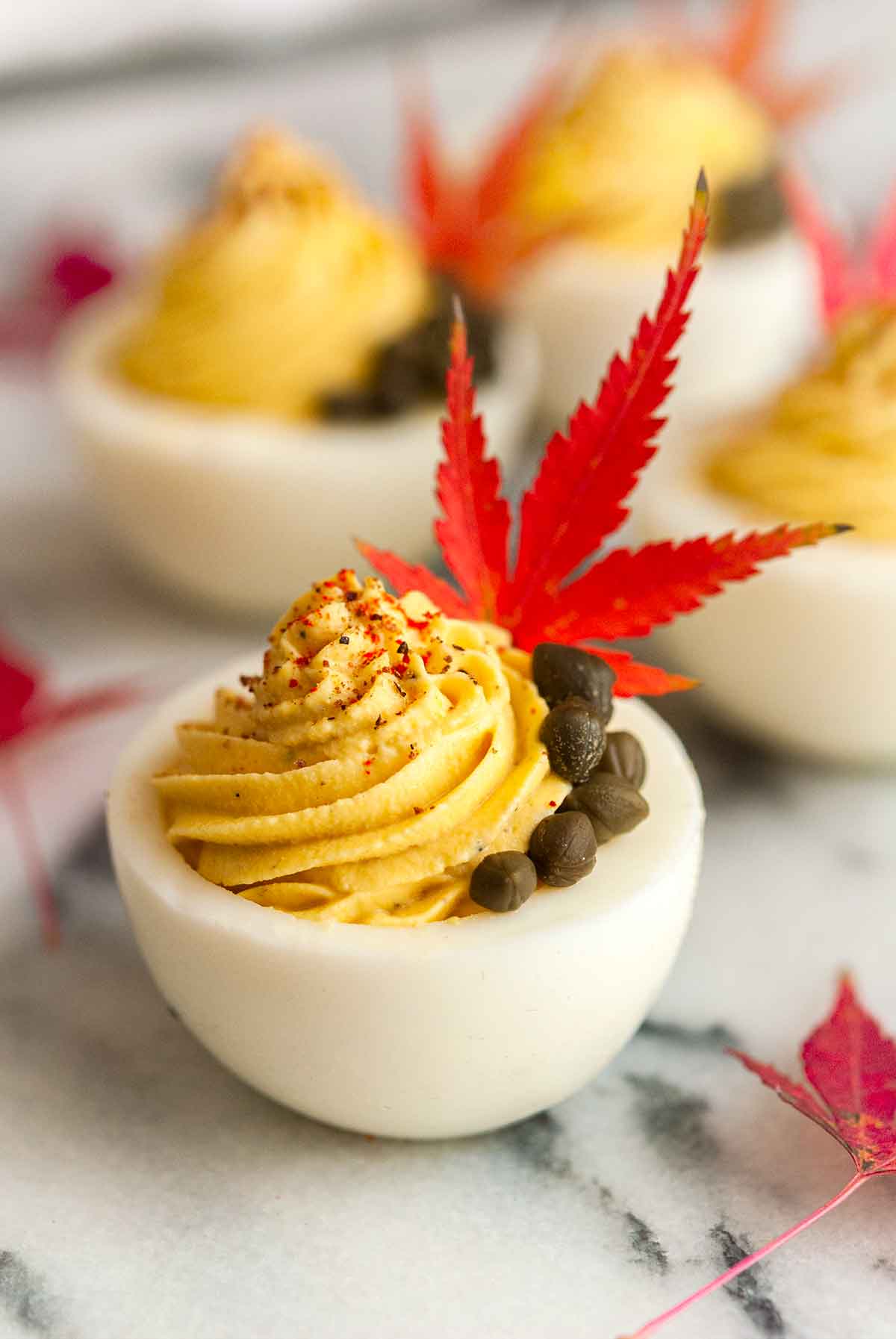A deviled egg garnished with capers, a single leaf, and sprinkle of spices in front of 3 others on a marble plate.