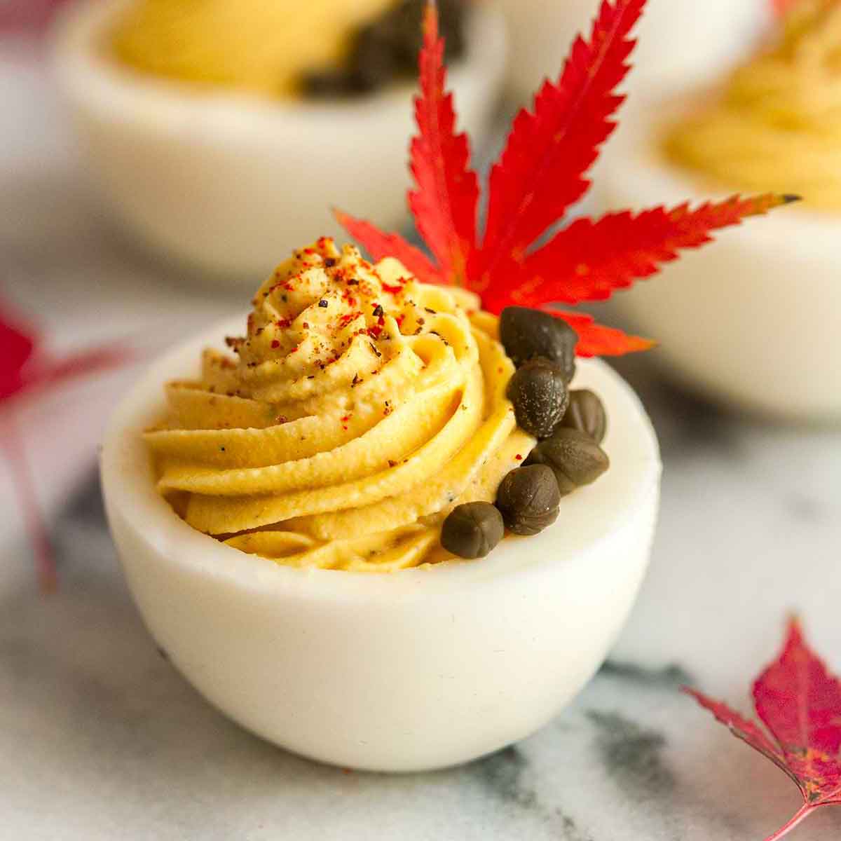 A deviled egg garnished with capers, a single leaf, and sprinkle of spices in front of 3 others on a marble plate.