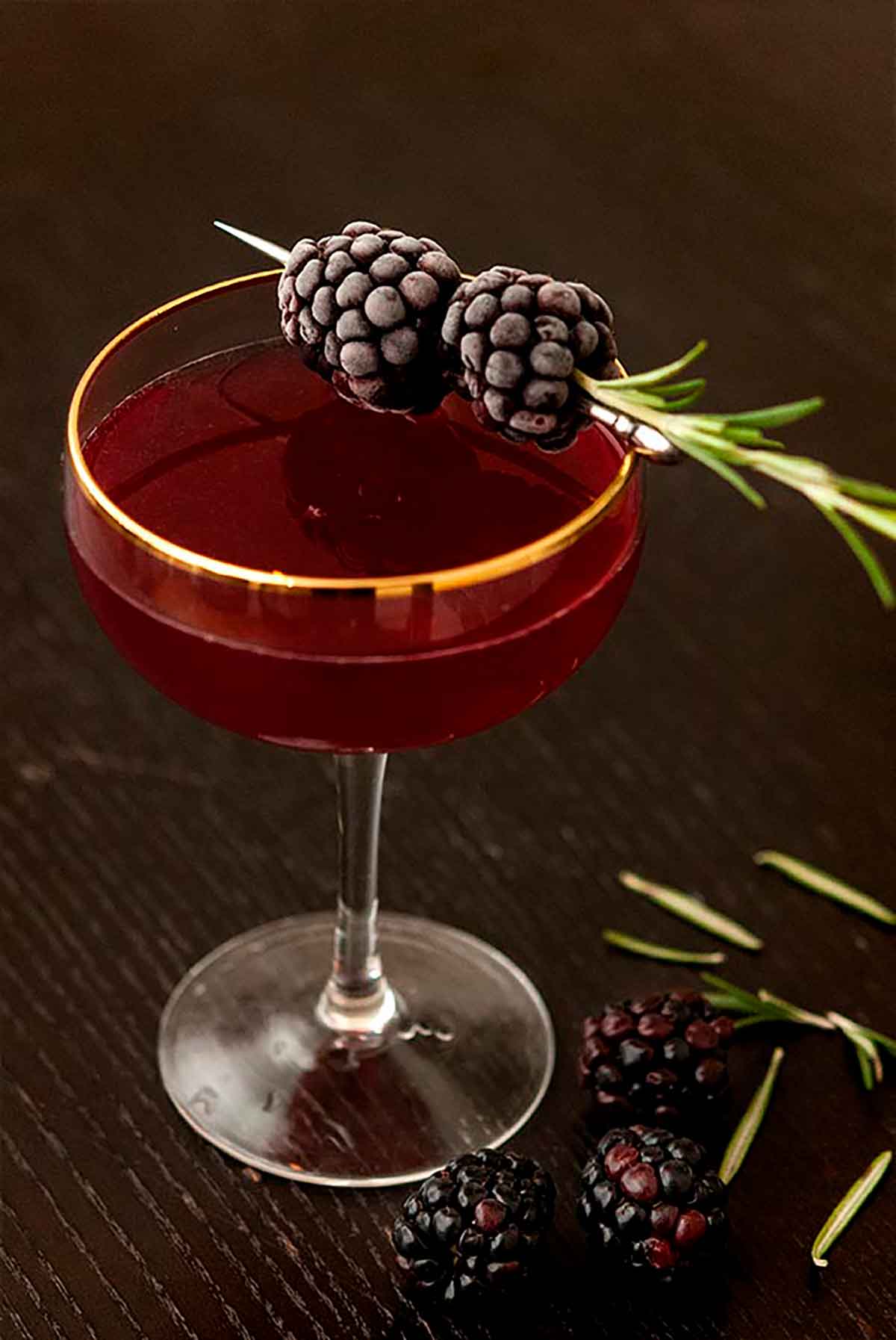 1 cocktail on a table surrounded by a few scattered blackberries, and garnished with frosted blackberries and rosemary.
