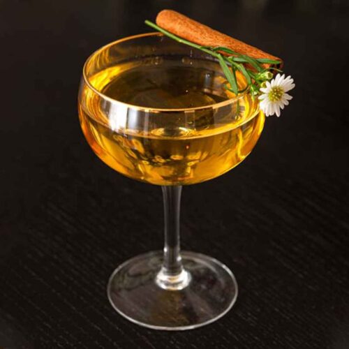 A cocktail with a gold rim on a black table, garnished with a cinnamon stick and daisy.