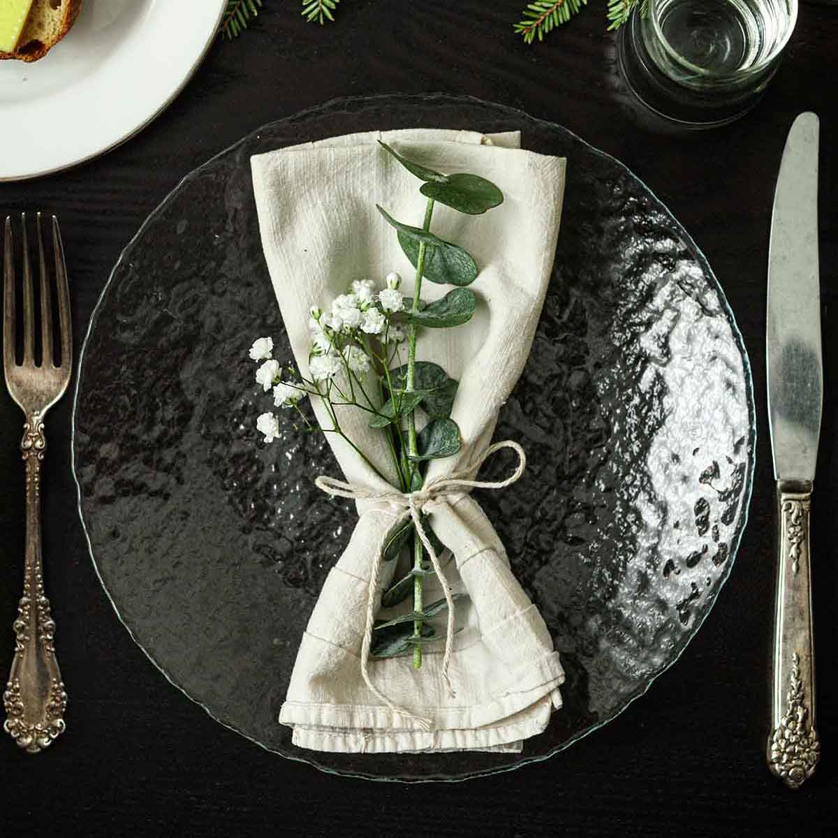 Eucalyptus and Baby’s Breath in a napkin on a table with holiday greenery, a plate of bread, silverware and glassware.