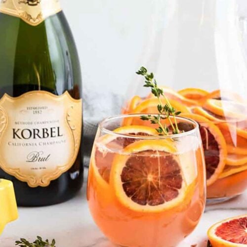 A cocktail filled with oranges and thyme beside a bottle of champagne on a marble table.