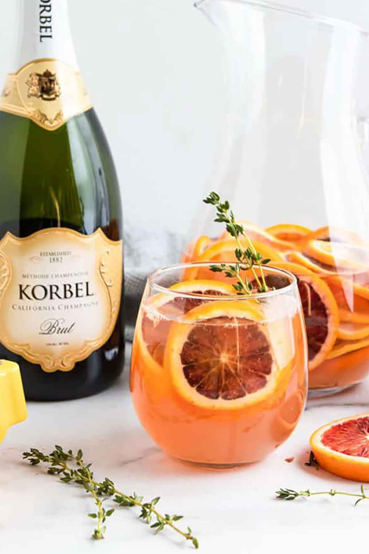 A cocktail filled with oranges and thyme beside a bottle of champagne on a marble table.