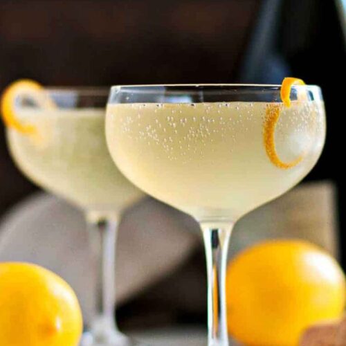 2 French 75 cocktails garnished with lemon on a table beside 2 lemons and a bottle in the background.