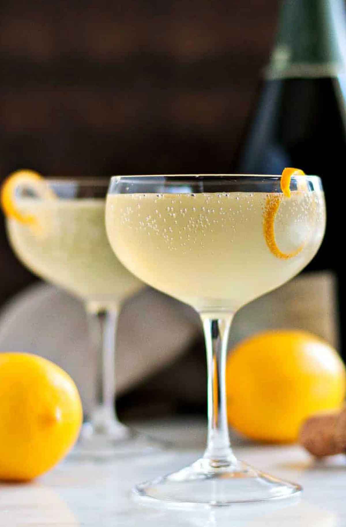 2 French 75 cocktails garnished with lemon on a table beside 2 lemons and a bottle in the background.