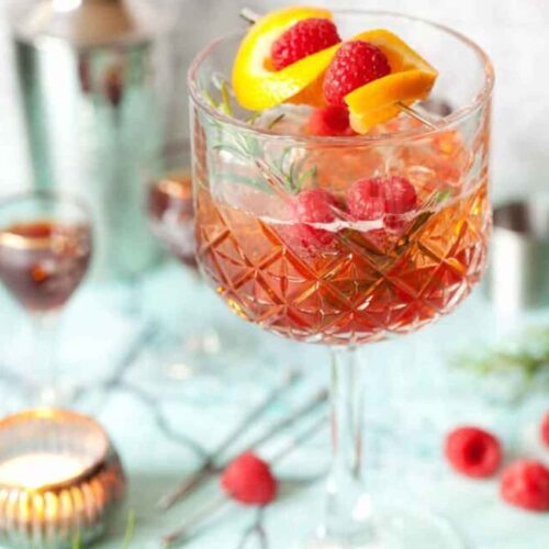 A cocktail on a table, sprinkled with raspberries, garnished with lemon and raspberries.