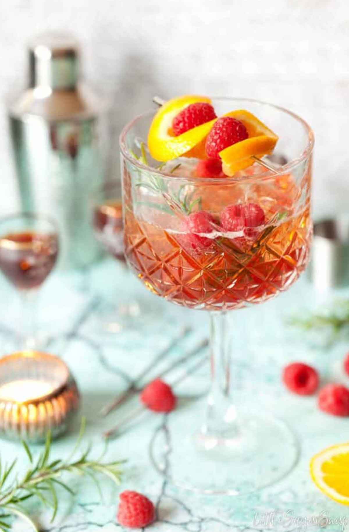 A cocktail on a table, sprinkled with raspberries, garnished with lemon and raspberries.