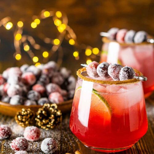 2 cocktails on a table with sugared cranberries and cinnamon sticks.