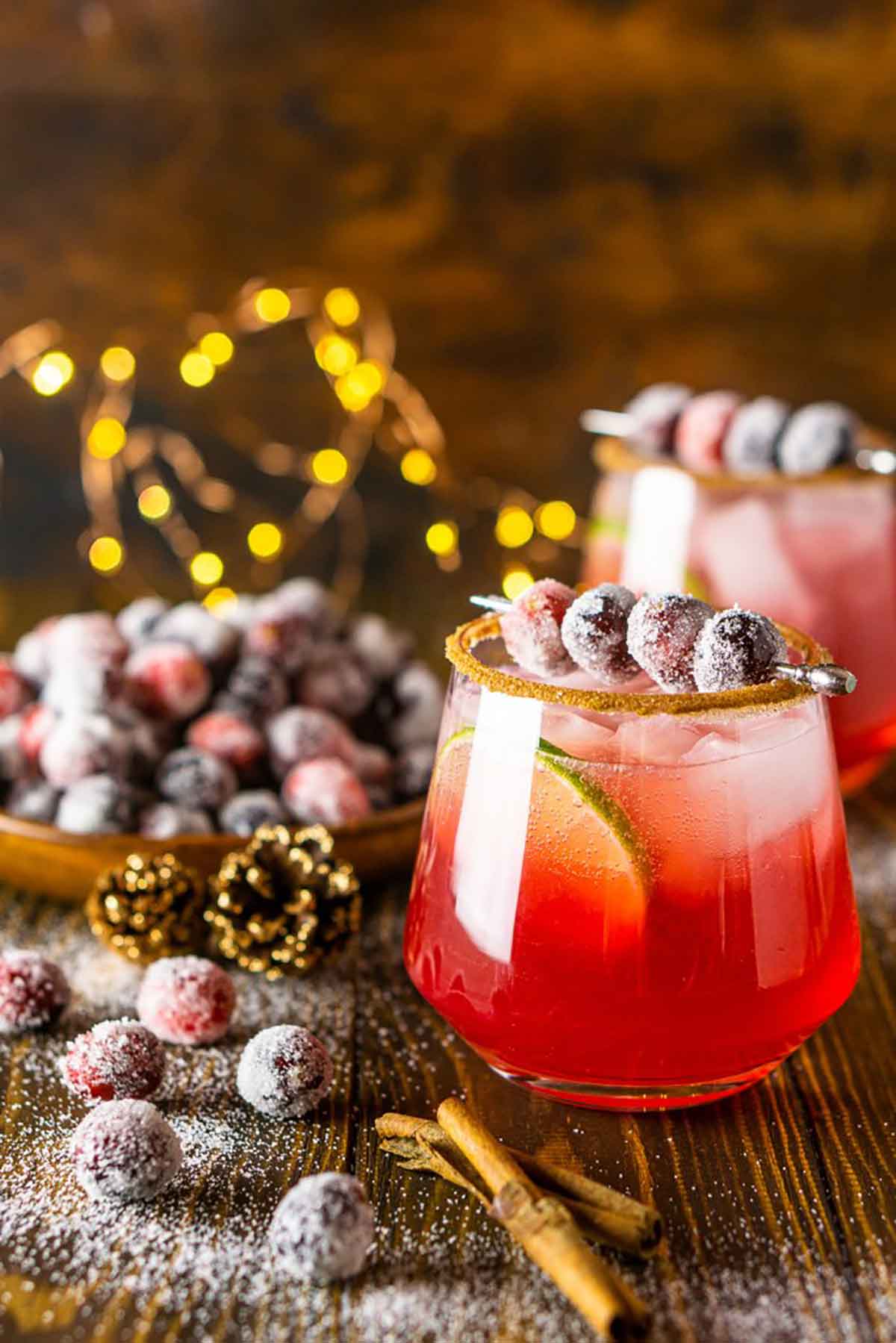 2 cocktails on a table with sugared cranberries and cinnamon sticks.