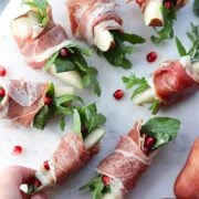 A hand reaching for 1 of 7 prosciutto-wrapped pears, garnished with pomegranate seeds on a plate.