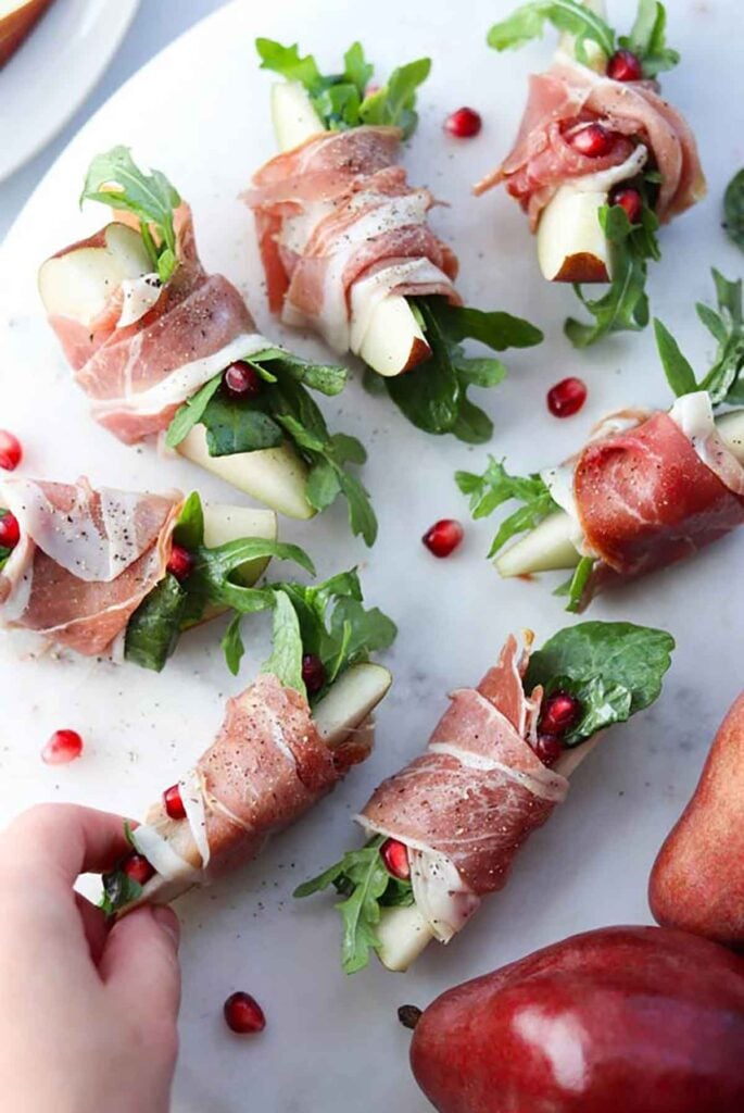 A hand reaching for 1 of 7 prosciutto-wrapped pears, garnished with pomegranate seeds on a plate.