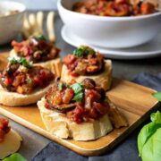 4 caponata crostini on a wooden board, in front of a bowl of caponata.