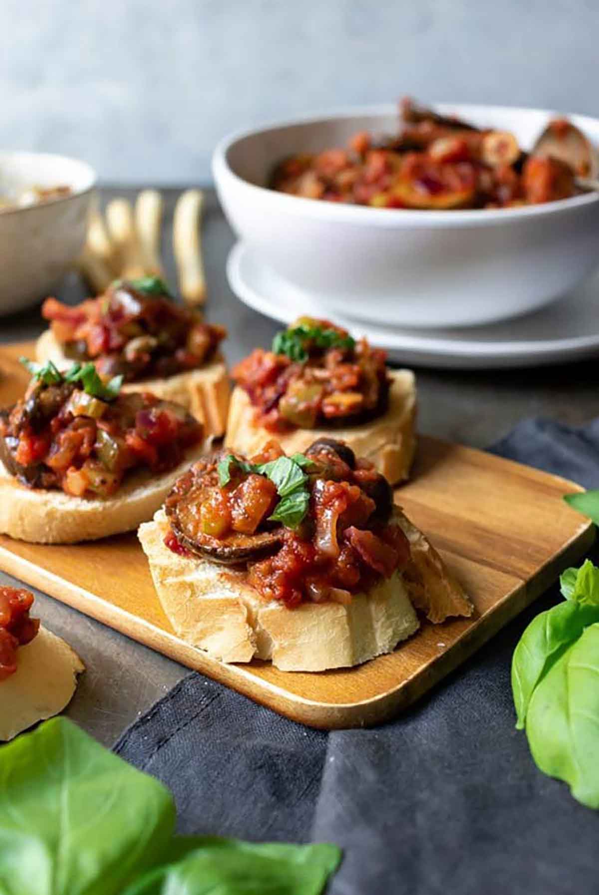 4 caponata crostini on a wooden board, in front of a bowl of caponata.