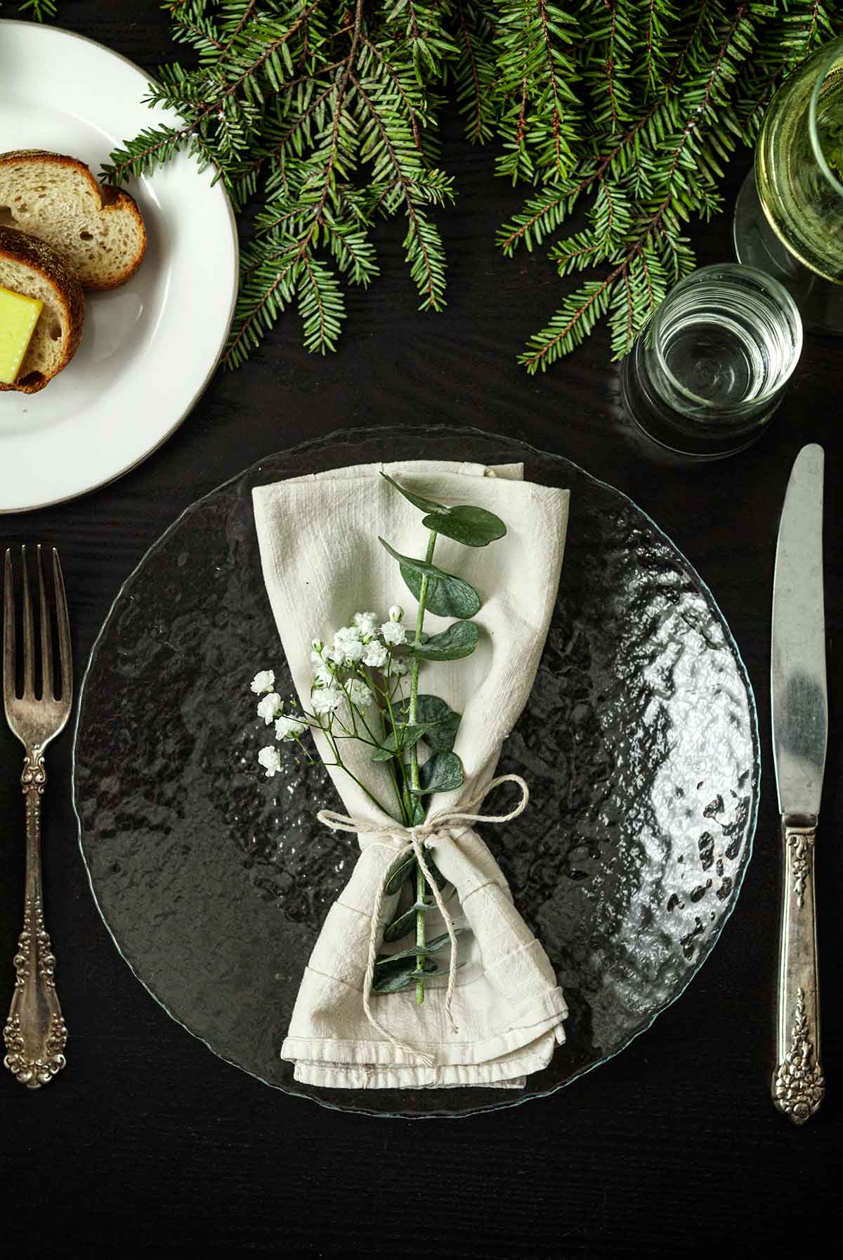 Eucalyptus and Baby’s Breath in a napkin on a table with holiday greenery, a plate of bread, silverware and glassware.