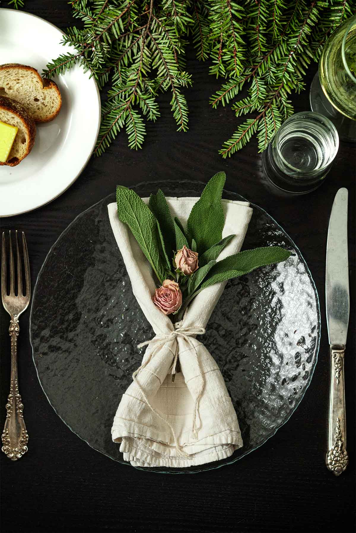 2 roses and sage in a tied napkin on a plate on a table with holiday greenery, a plate of bread, silverware and glassware.