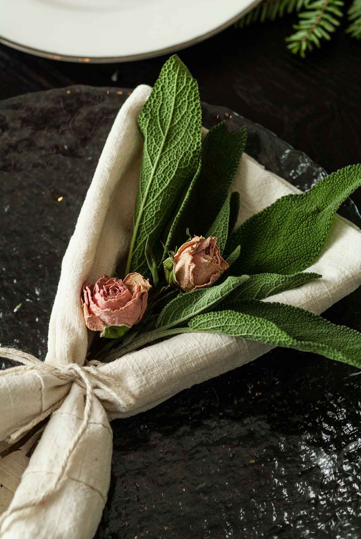 2 small roses and a few leaves of sage in a napkin tied with string, on a plate.