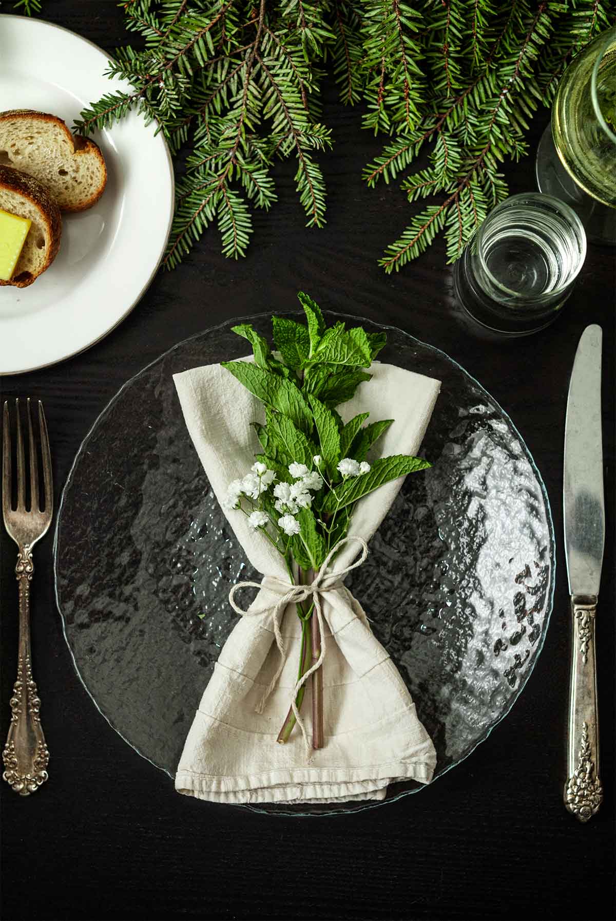 Mint and Baby’s Breath in a napkin on a plate on a table with holiday greenery, a plate of bread, silverware and glassware.