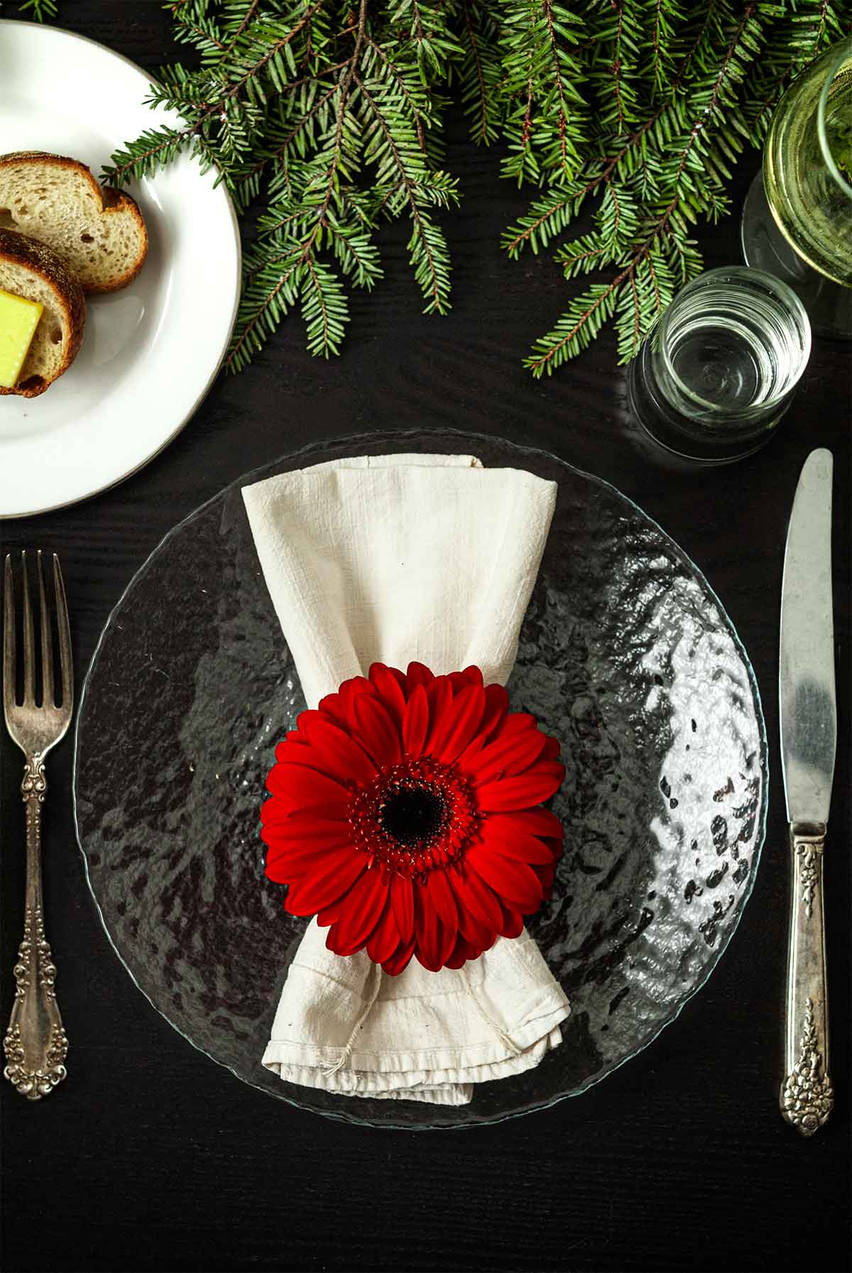 A Gerbera Daisy on top of a napkin on a plate on a table with holiday greenery, a plate of bread, silverware and glassware.