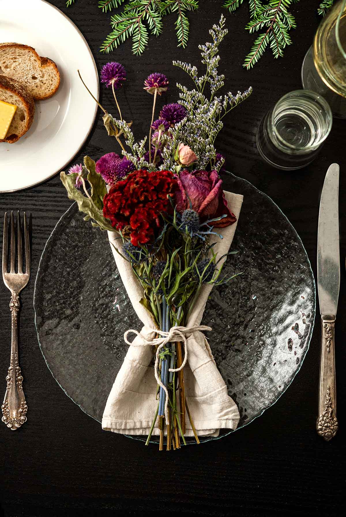 A bouquet of flowers in a napkin on a plate on a table with holiday greenery, a plate of bread, silverware and glassware.