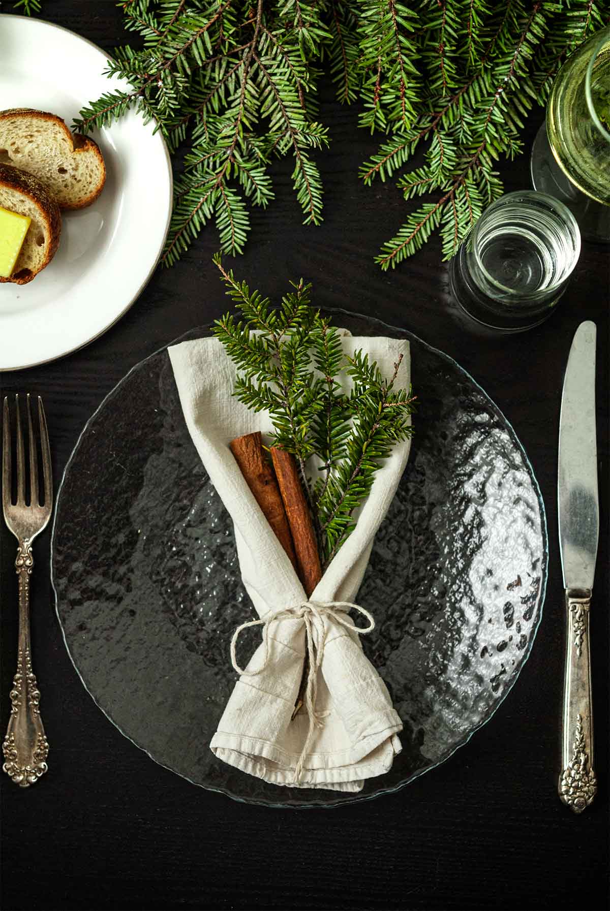 Sprigs of pine and cinnamon sticks in a napkin on a plate on a table with greenery, a plate of bread, silverware and glasses.