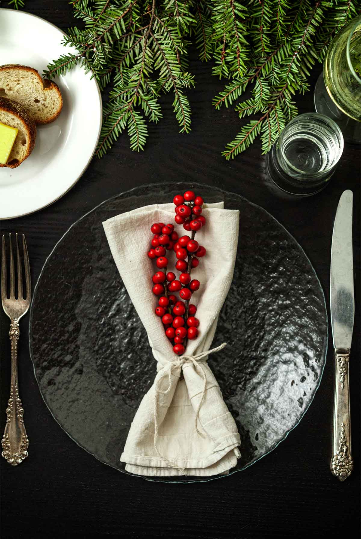 2 sticks of Ilex in a tied napkin on a plate on a table with holiday greenery, a plate of bread, silverware and glassware.