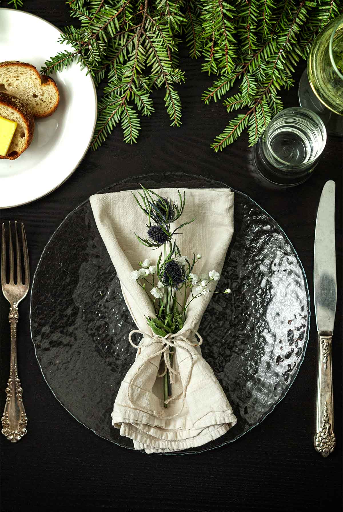 Thistle and Baby’s Breath in a napkin on a plate on a table with greenery, a plate of bread, silverware and glassware.