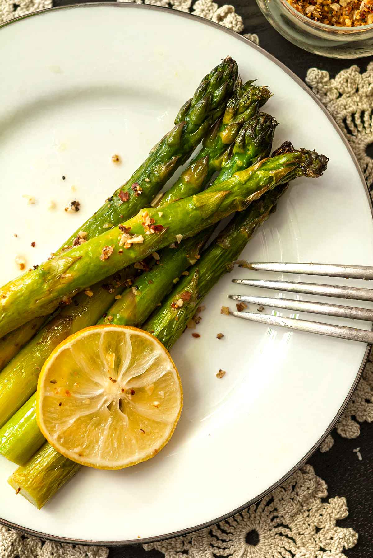 5 asparagus stalks on a plate with a fork, sprinkled with lemon pepper and garnished with a lemon slice.