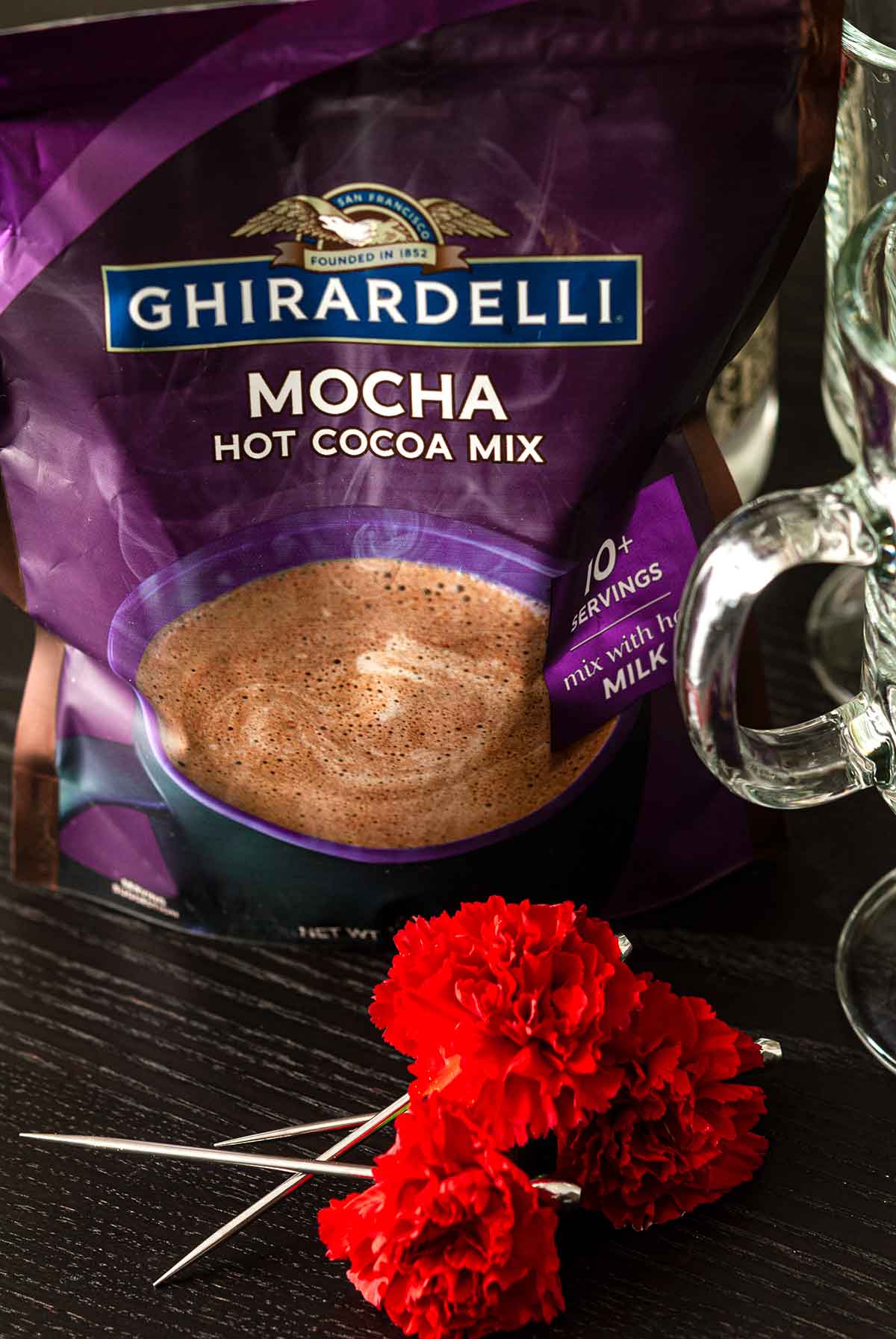 A bag of Ghiradelli mocha hot cocoa mix on a table behind 3 flower garnishes.