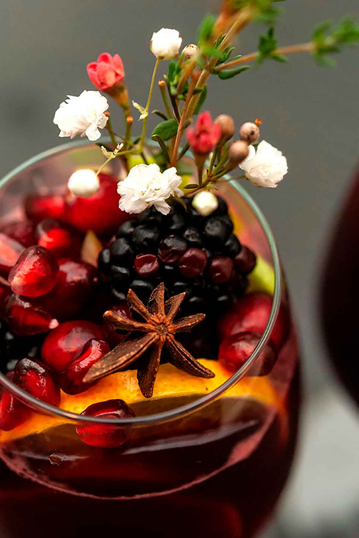 A glass of sangria garnished with fruits, flowers, herbs and star anise on a dark table.