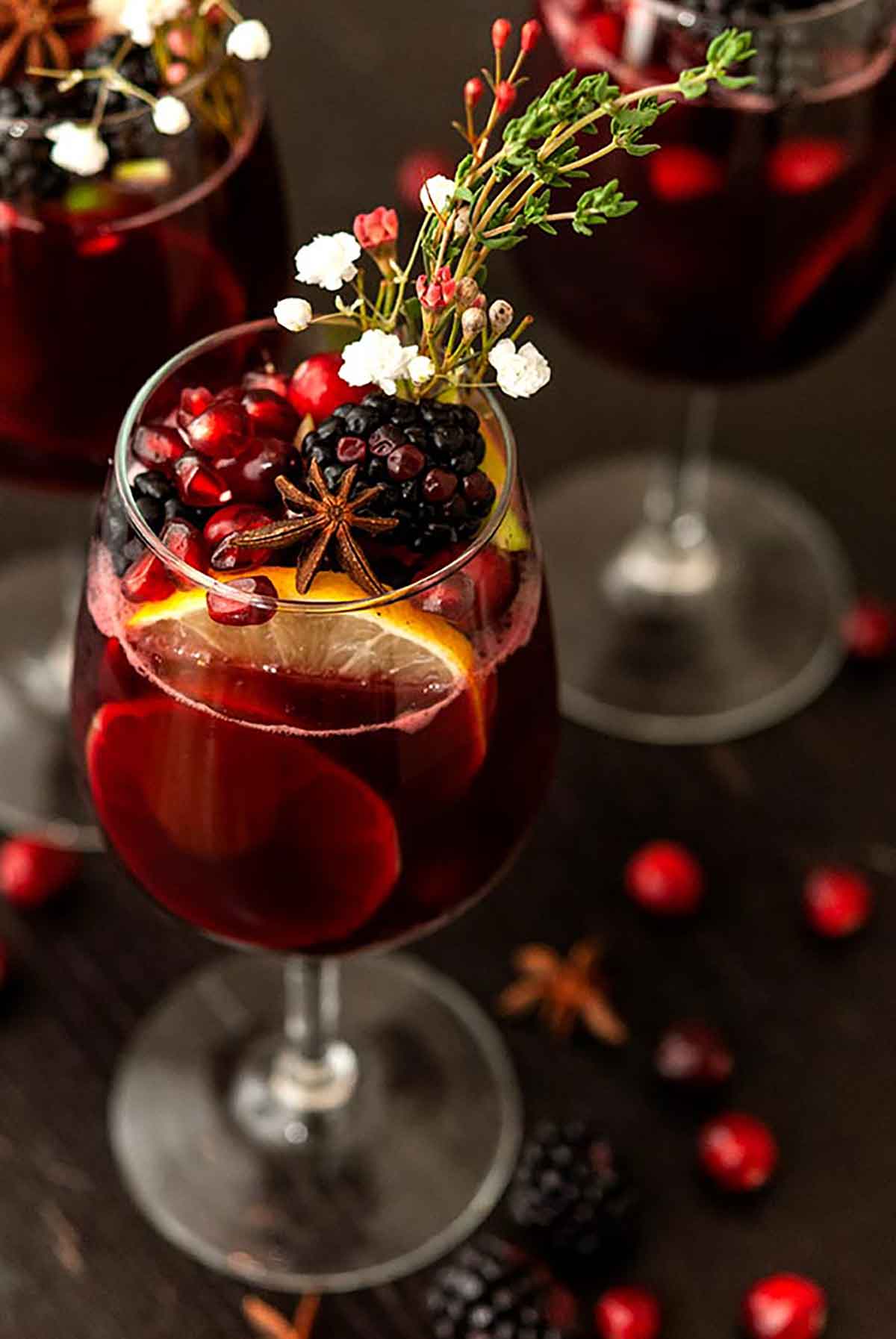 A glass of sangria garnished with fruits, flowers, herbs and star anise on a dark table.