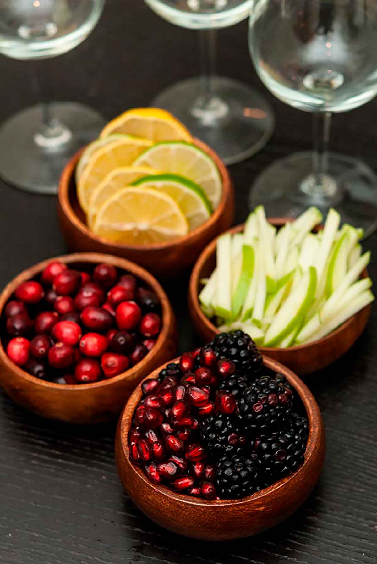 Berries, citrus and apple garnishes in small wooden bowls on a table with 3 empty wine glasses.